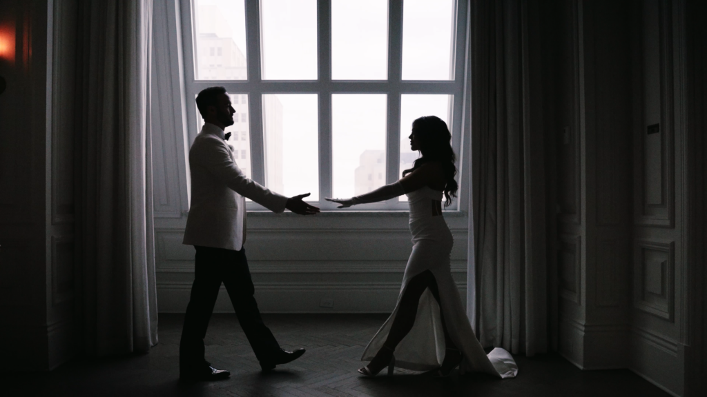 Bride and groom walking toward each other while reaching out in front of a large window in this hotel space in Dallas.