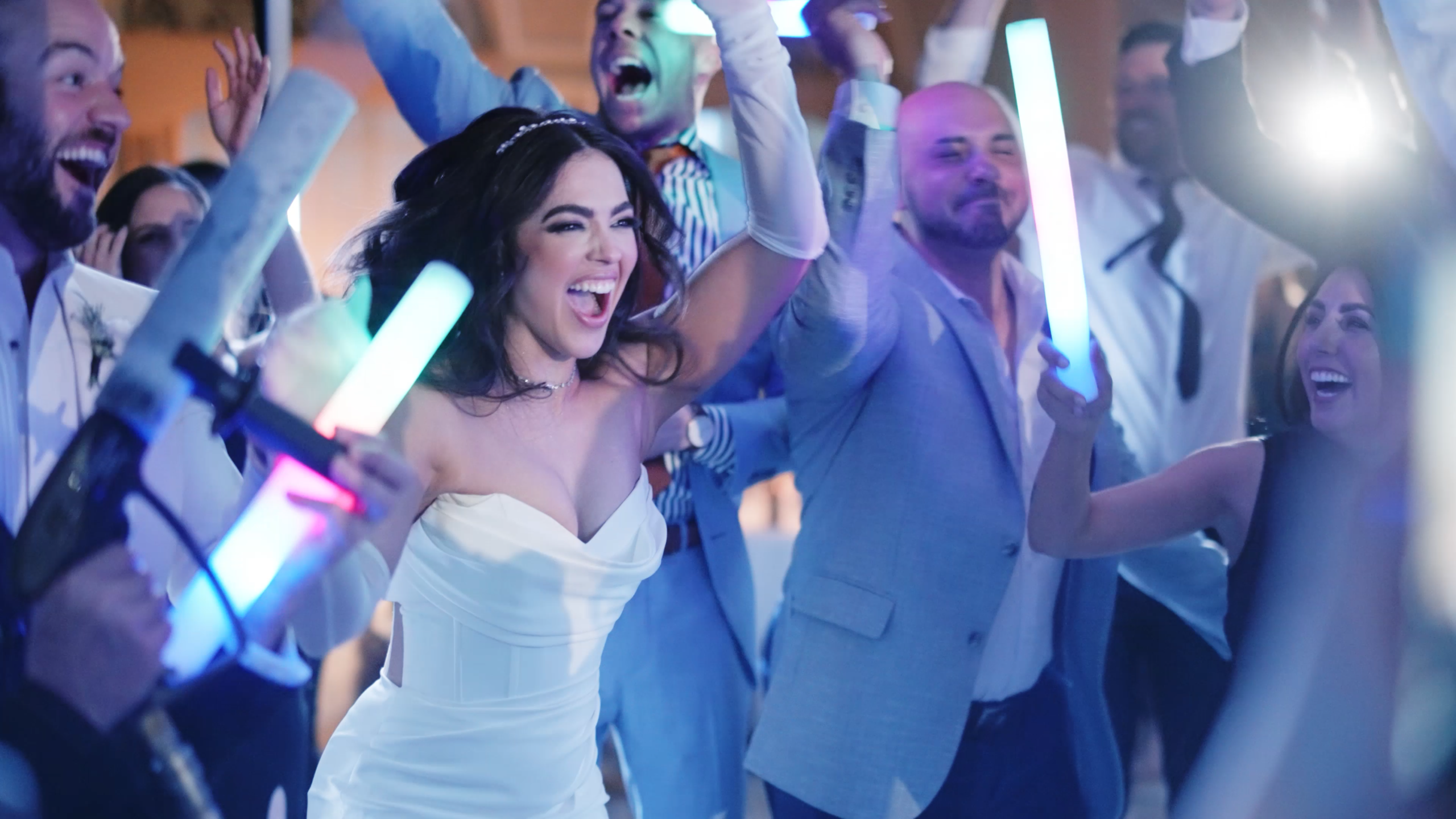 Bride at her wedding reception laughing joyfully with her guests while holding a glowing light stick, surrounded by a lively, celebratory atmosphere.