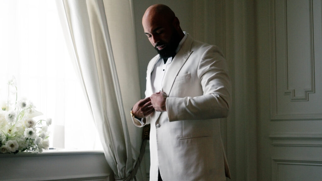 Groom looking down as he buttons his jacket while getting ready for his wedding next to a large window.
