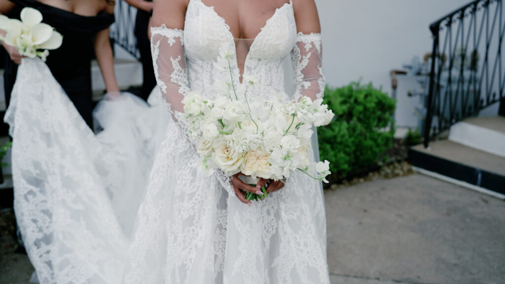 Bride shown from neck to knees, wearing her wedding dress, with a bridesmaid standing behind her; both are captured from behind or partially turned away so their faces are not visible.