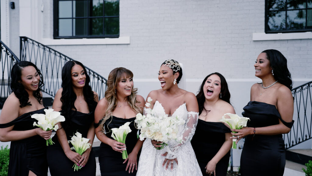 Bride laughing with her bridesmaids who are in all black, as they stand outside her Dallas wedding venue.