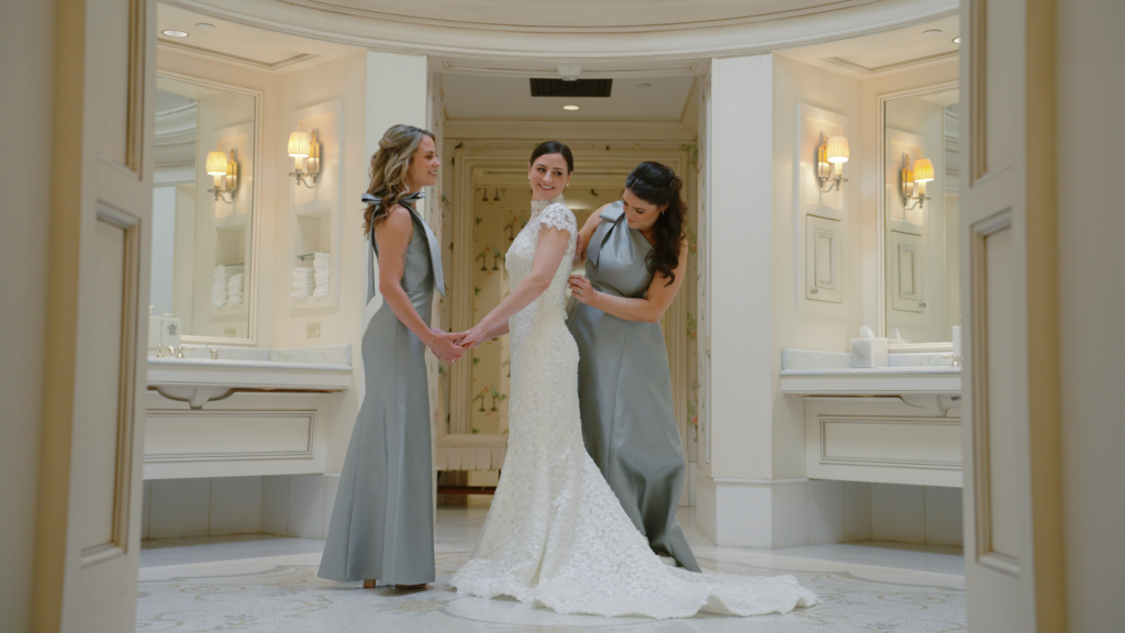 Bridesmaids assist the bride with her finishing touches of getting ready, while one holds her hands and the other buttons her wedding dress. They are in a beautiful white room with large mirrors.