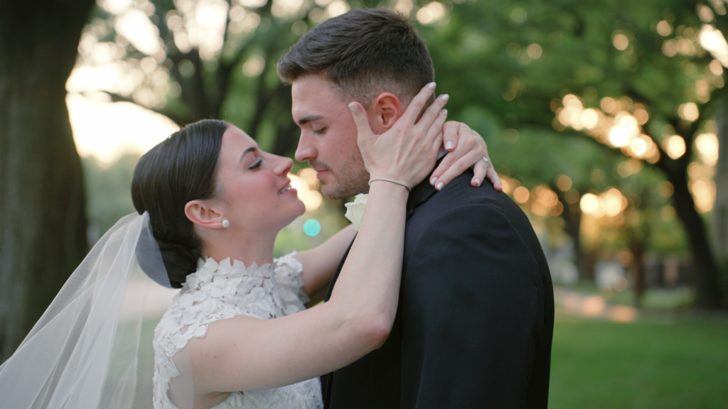 Bride and groom share a romantic moment, leaning in for a kiss, prior to their wedding ceremony as trees surround them and the sun goes down in the background.