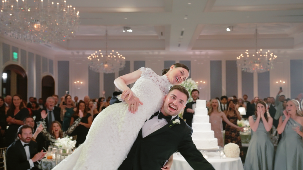 Groom picking up his bride and holding her on his shoulder at their wedding reception, with guests looking on and cheering.