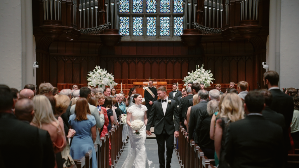 A bride and groom exit their ceremony hand-in-hand down the aisle as guests stand and clap at their Dallas wedding.