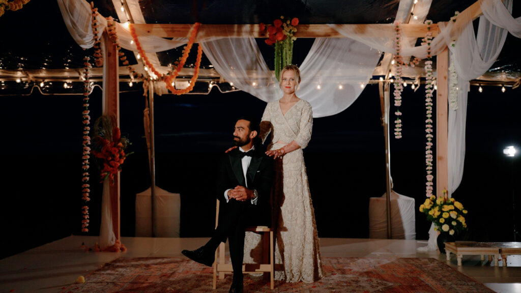 Bride and groom outside in their New England wedding ceremony set up with the bride standing and her hands on her groom's shoulders while he sits.