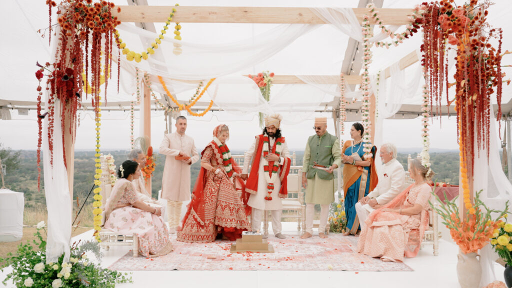 An elaborate Indian wedding with family members sitting and watching the ceremony take place underneath colorful beading and florals surrounding them.