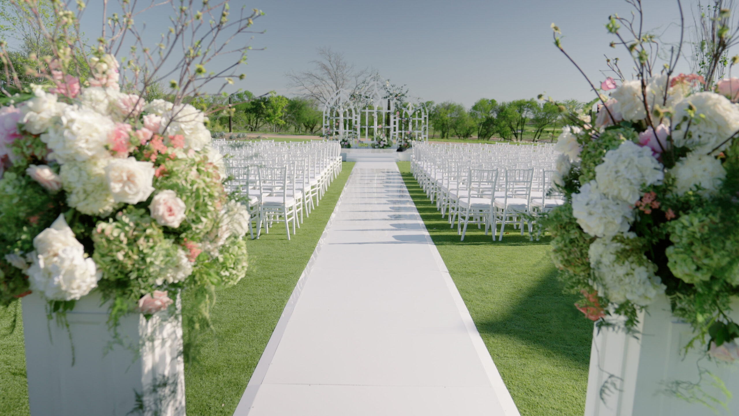 A wedding aisle that leads to a beautiful backdrop outside, with white chairs and flowers lining the area.