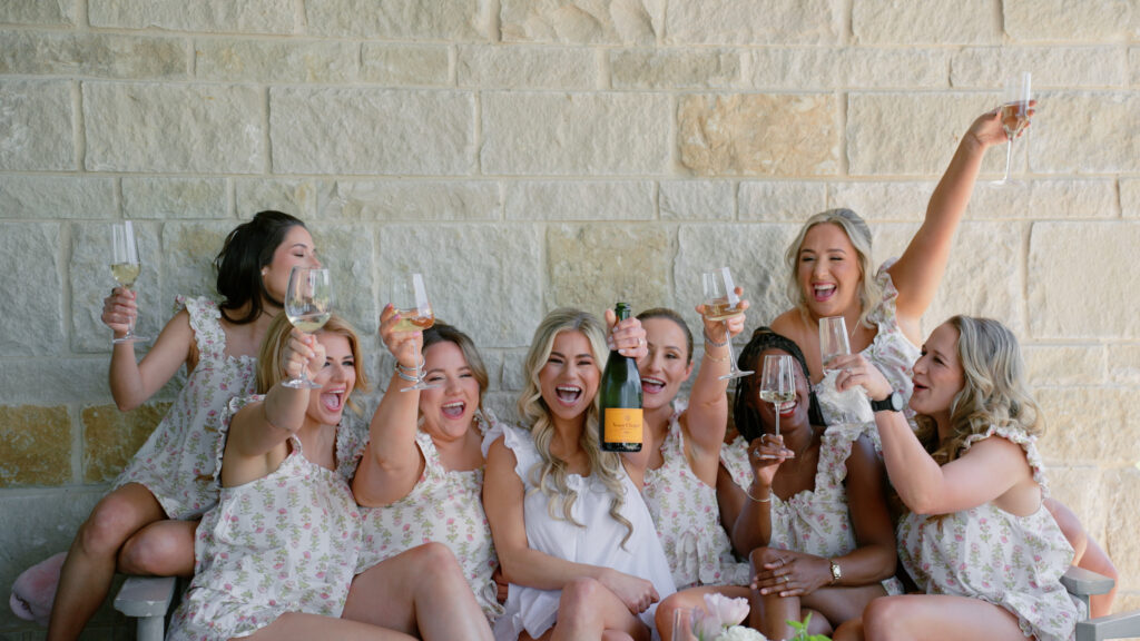 Bride and bridesmaids toasting with their champagne, while the bridesmaids hold up their glasses and the bride holds up the whole bottle. All smiling and enjoying the wedding day in Dallas, TX.