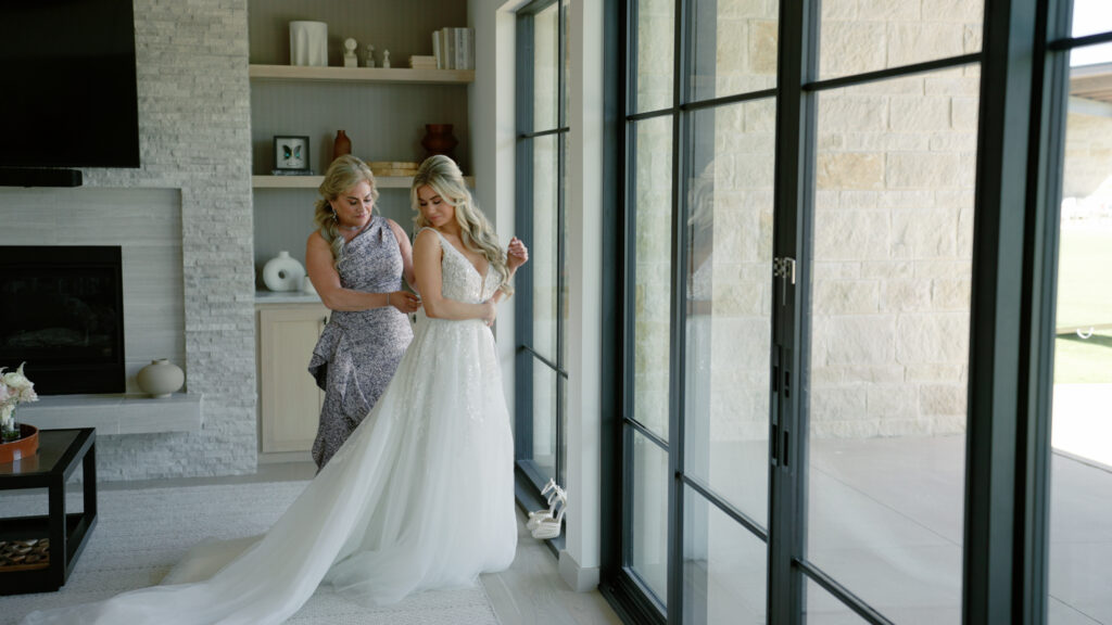 Mother buttoning her daughter’s wedding gown in a hotel room during a quiet, emotional getting-ready moment.