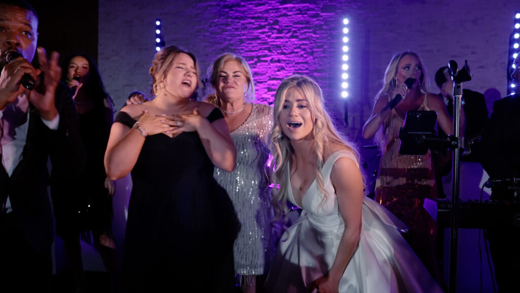 A bride, her bridesmaid, and her mother, all stand on stage singing a song at her reception on her wedding night in Boston, MA.