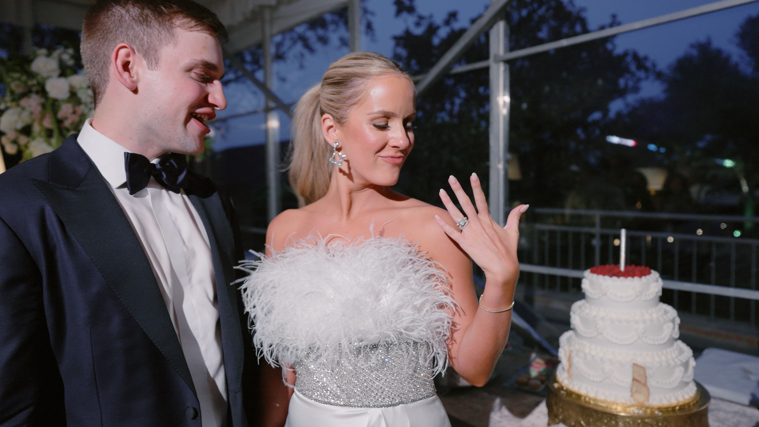 A bride flashes her ring and looks down at it while her groom stands smiling next to her with their wedding cake in the background.