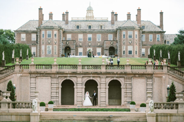 Couple posing for a wedding photo in front of Castle Hill on the Crane Estate, standing together on the manicured grounds with the historic mansion and sweeping coastal landscape in the background.