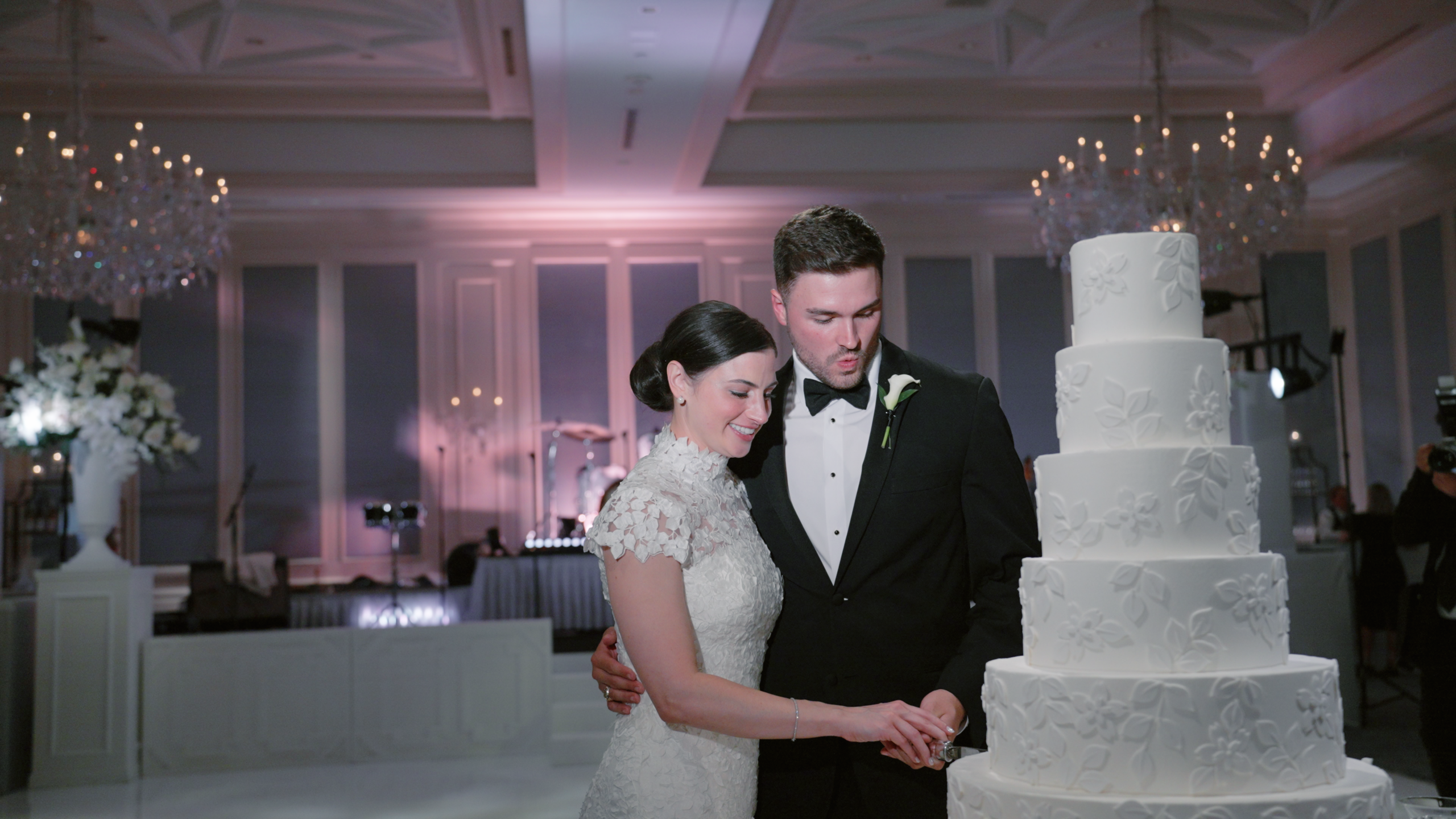 Bride and groom cutting their wedding cake together, smiling during a joyful reception moment surrounded by guests.
