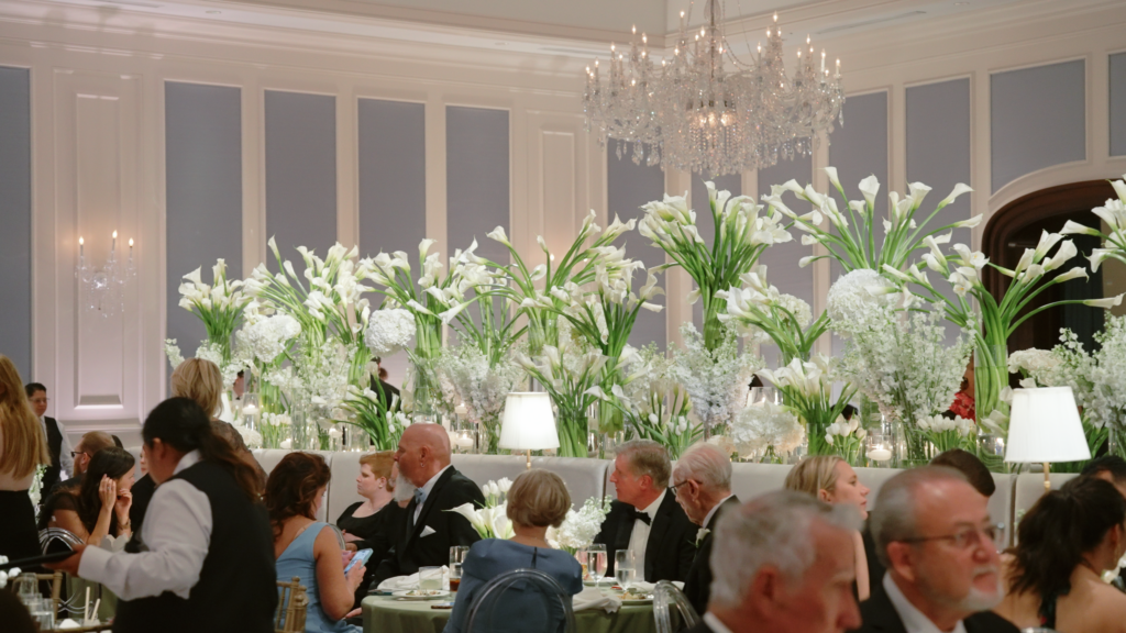 Elegant indoor wedding reception with blue paneled walls, guests seated at tables, and large white floral centerpieces.