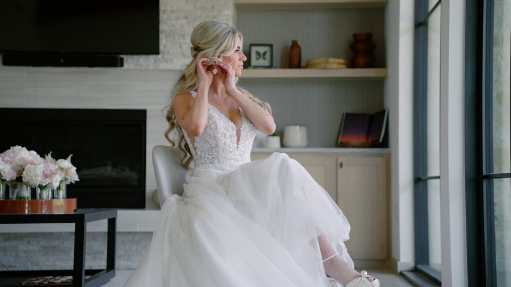 Bride putting on earrings while looking out the window during wedding morning preparations.