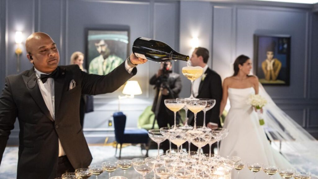Man pouring champagne into a cascading tower of coupe glasses at The Newbury Boston while the bride and groom stand nearby watching and smiling during their wedding reception.